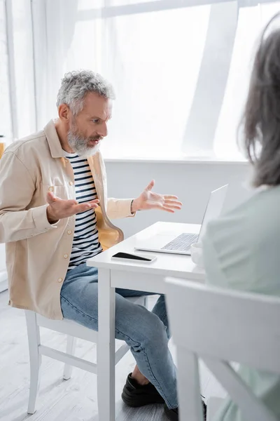 Middle aged freelancer having video call on laptop near blurred wife at home — Stock Photo