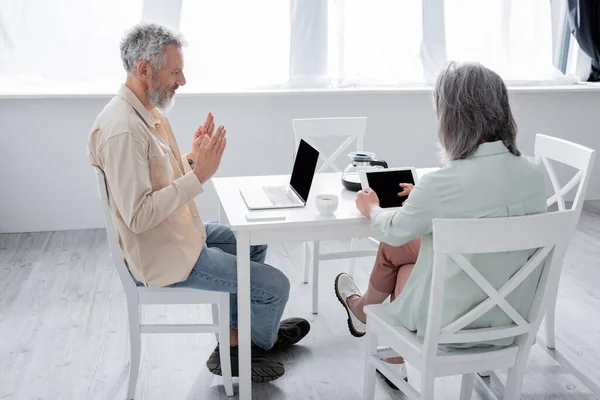 Homme mature montrant un geste correct lors d'un appel vidéo sur un ordinateur portable près de la femme avec une tablette numérique à la maison — Photo de stock