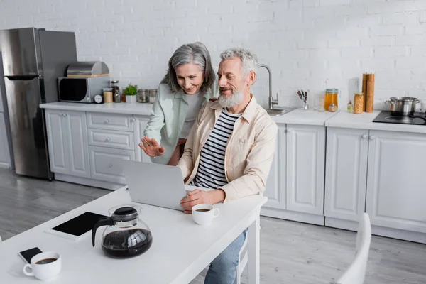 Coppie mature sorridenti che fanno videochiamata su computer portatile vicino a dispositivi e caffè in cucina — Foto stock