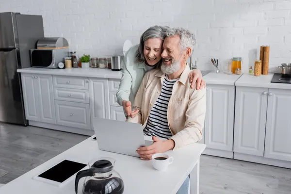 Mujer madura alegre abrazando marido y apuntando a la computadora portátil cerca de café y dispositivos en casa - foto de stock
