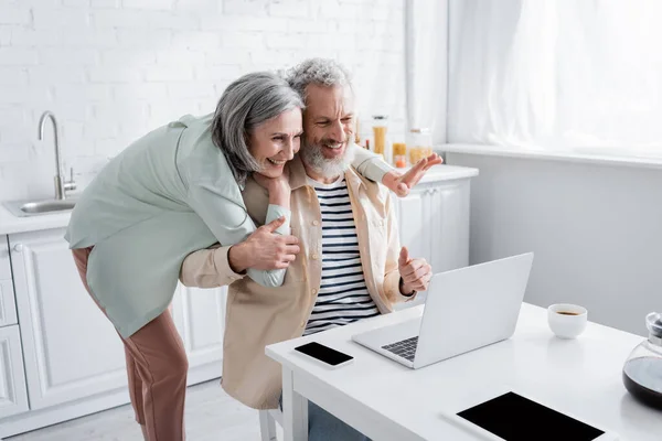 Couple mature gesticulant lors d'un appel vidéo sur un ordinateur portable près du café dans la cuisine — Photo de stock