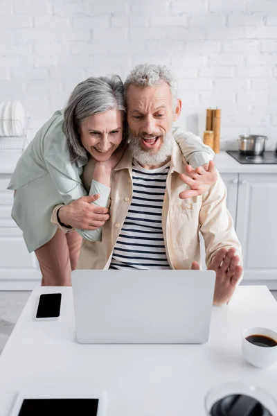 Cheerful middle aged couple having video call on laptop near coffee and devices in kitchen — Stock Photo