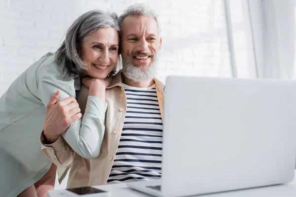 Positive reife Frau umarmt Ehemann und schaut auf verschwommenen Laptop zu Hause — Stockfoto