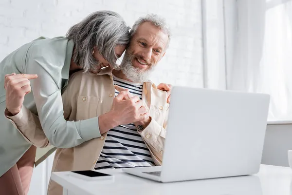 Lächelnder Mann zeigt bei Videoanruf am heimischen Laptop auf Ehefrau — Stockfoto