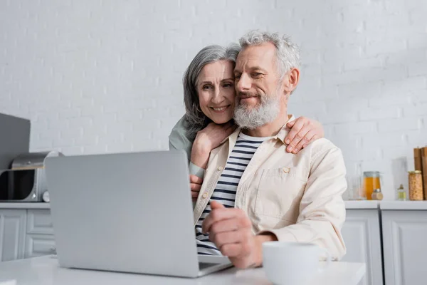 Smiling mature woman hugging husband near blurred laptop and cup in kitchen — Stock Photo