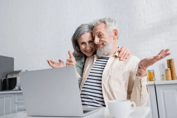 Mujer positiva abrazando marido confundido cerca del ordenador portátil y taza de café en la cocina - foto de stock
