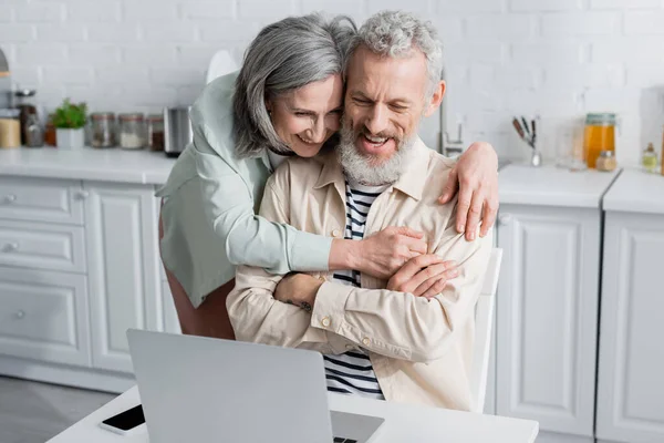 Mature woman hugging cheerful husband looking at laptop near smartphone in kitchen — Stock Photo