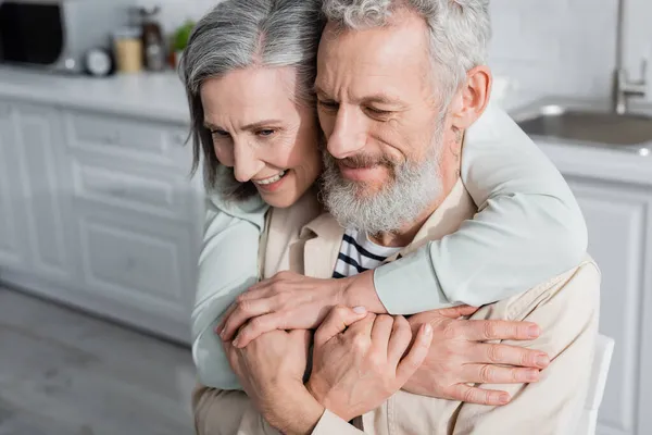 Smiling woman hugging mature husband in kitchen — Stock Photo