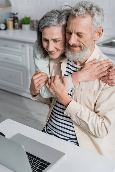 Mature couple hugging near laptop in kitchen at home — Stock Photo