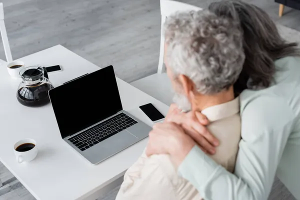 Devices and coffee on table near blurred mature woman hugging husband at home — Stock Photo
