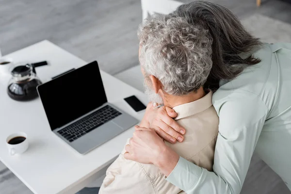 Femme d'âge moyen embrassant mari près de gadgets et café à la maison — Photo de stock