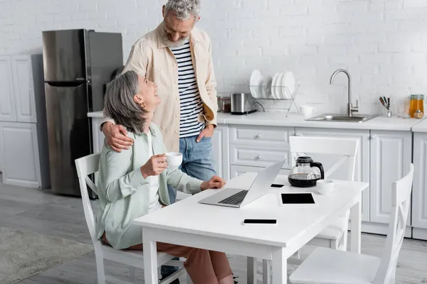 Homme joyeux étreignant femme avec du café près de gadgets dans la cuisine — Photo de stock