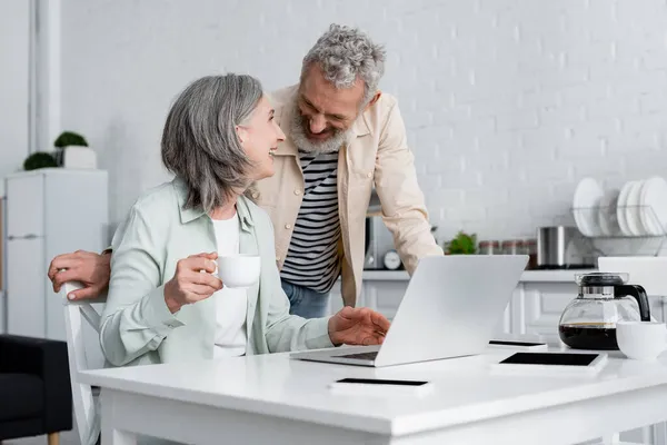 Pareja madura positiva hablando cerca de café y gadgets en la cocina - foto de stock