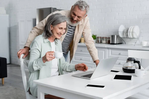 Mature freelancer with coffee using laptop near husband in kitchen — Stock Photo