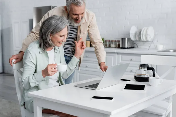 Positive woman holding cup and pointing at husband during video call on laptop at home — Stock Photo