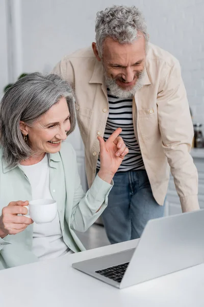 Femme souriante avec café pointant vers mari mature pendant le chat vidéo sur ordinateur portable dans la cuisine — Stock Photo