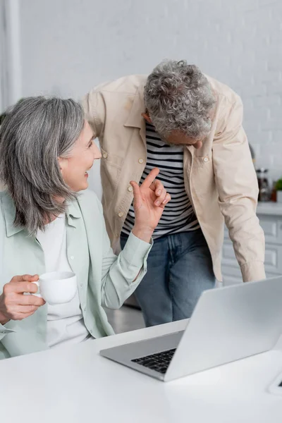 Mujer de mediana edad sosteniendo la taza y señalando al marido cerca de la computadora portátil borrosa en la mesa de la cocina - foto de stock