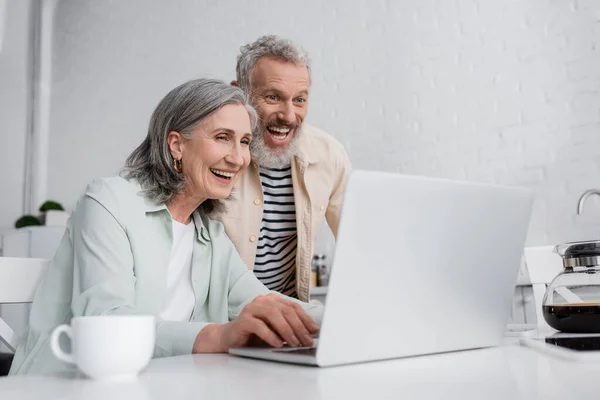 Positive mature couple looking at blurred laptop near coffee pot in kitchen — Stock Photo