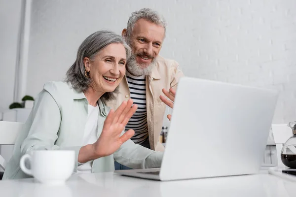 Mature couple having video chat on laptop near blurred cup of coffee at home — Stock Photo