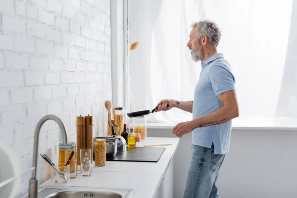 Seitenansicht des reifen Mannes beim Kochen von Pfannkuchen in der Küche. Übersetzung: 