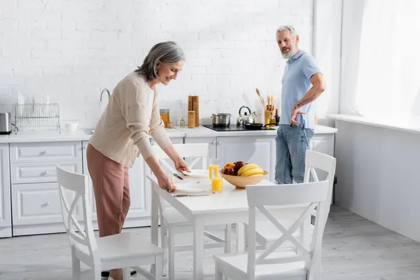 Mujer sonriente poniendo platos en la mesa cerca del jugo de naranja, frutas y marido en la cocina - foto de stock