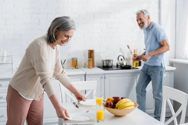 Cheerful mature woman putting plates near orange juice ad blurred husband in kitchen — Stock Photo