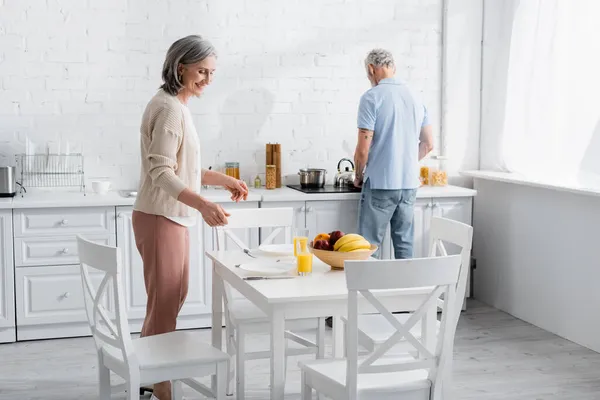 Reife Frau steht neben Orangensaft und Früchten auf Tisch in Küche — Stockfoto