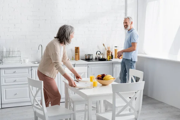 Uomo sorridente che guarda moglie vicino a succo d'arancia e frutta sul tavolo in cucina in cucina — Foto stock