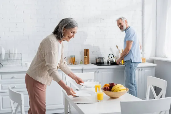 Mulher sorridente segurando pratos perto da mesa e marido borrado cozinhar na cozinha — Fotografia de Stock