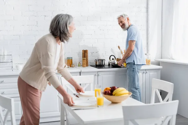 Positivo hombre maduro cocina y mirando a la esposa con platos en la cocina - foto de stock