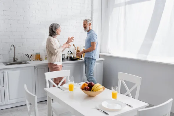 Vue latérale du couple d'âge mûr souriant parlant pendant la cuisson dans la cuisine — Photo de stock