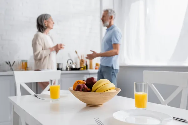 Frutas e suco de laranja na mesa perto de casal maduro desfocado na cozinha — Fotografia de Stock