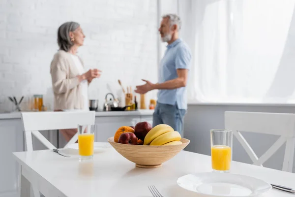 Frutas y zumo de naranja en la mesa cerca de pareja madura borrosa en la cocina - foto de stock