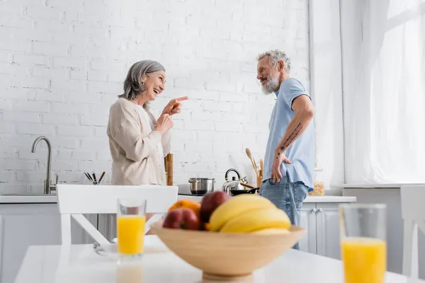 Donna sorridente che punta al marito vicino stufa e frutta sfocata in cucina — Foto stock