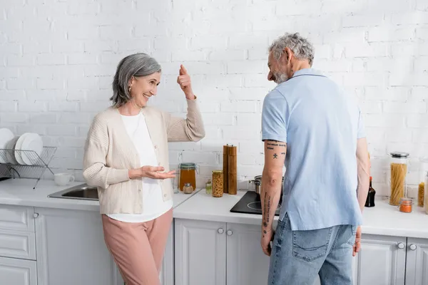 Woman pointing near husband in kitchen. Translation: 