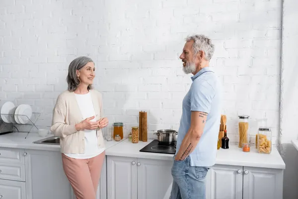 Donna sorridente che parla con marito vicino stufa e piano di lavoro in cucina — Foto stock