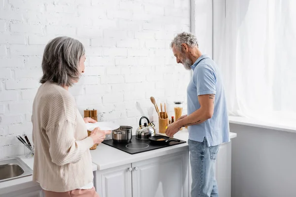 Maduro hombre cocina panqueque cerca de la esposa con plato en la cocina - foto de stock