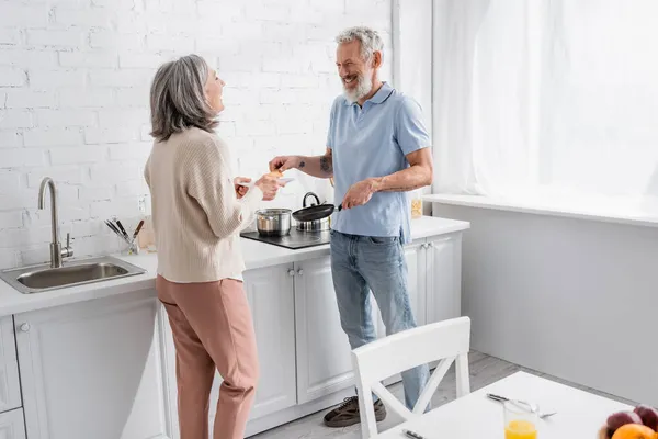 Glücklich reifer Mann mit Pfanne und Pfannkuchen in der Nähe seiner Frau mit Teller in der Küche — Stockfoto