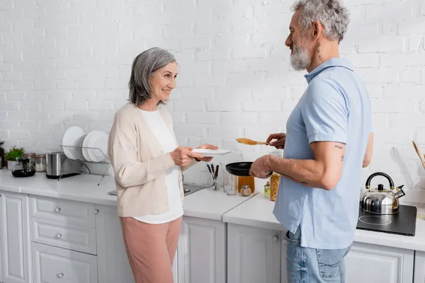 Woman holding plate near husband with pancake and frying pan in kitchen — Stock Photo