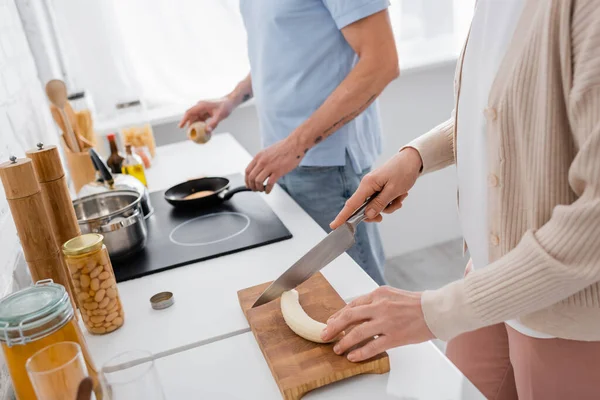 Cropped view of woman cutting banana near blurred husband cooking in kitchen — Stock Photo