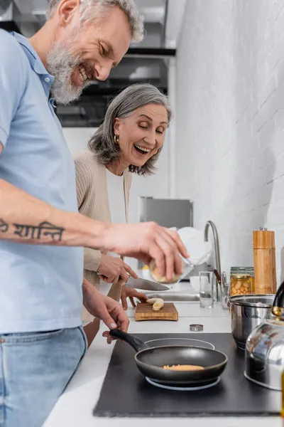 Mujer sosteniendo cuchillo cerca de marido cocinando panqueques. Traducción: 