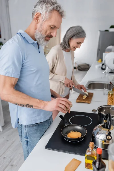 Side view of man seasoning pancake near blurred wife cutting banana in kitchen — Stock Photo