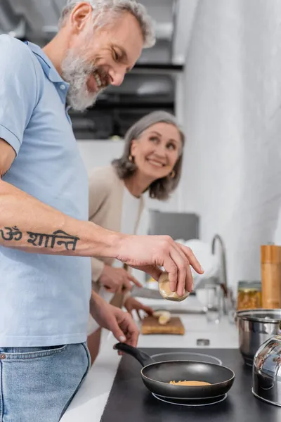 Homme souriant tenant épice tout en cuisinant une crêpe près de femme floue dans la cuisine — Photo de stock