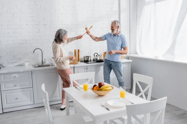 Side view of smiling mature couple fighting with spatulas in kitchen — Stock Photo