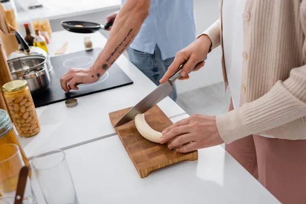 Vista recortada de la mujer cortando plátano cerca de marido borroso con sartén en la cocina - foto de stock