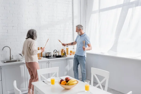 Cheerful mature couple holding spatulas near worktop in kitchen — Stock Photo