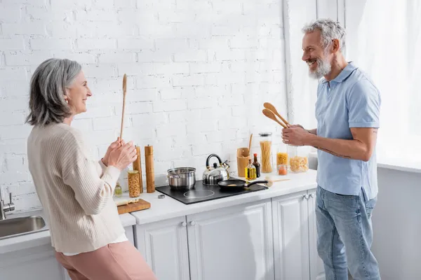 Felice coppia matura tenendo cucchiai vicino stufa in cucina — Foto stock