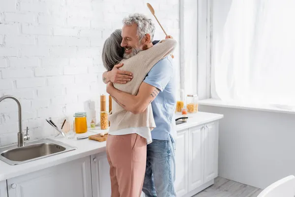 Uomo sorridente che abbraccia moglie con cucchiaio in cucina — Foto stock