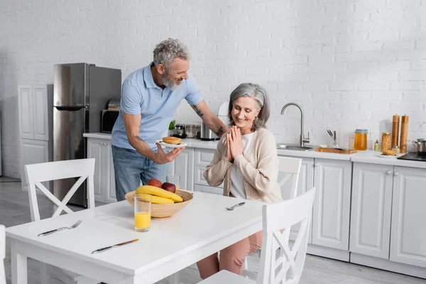 Alegre uma exploração panquecas perto de esposa e frutas na cozinha — Fotografia de Stock