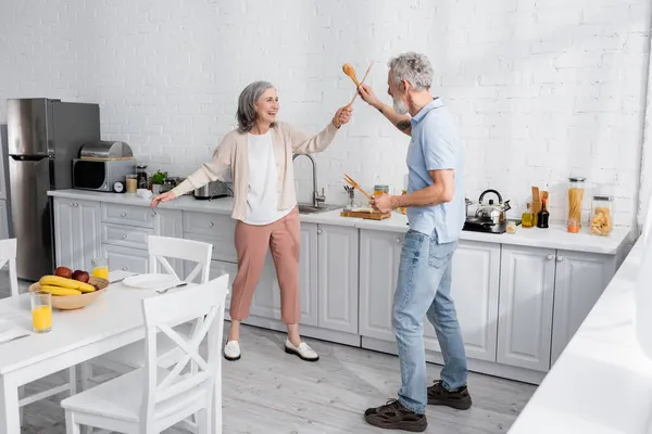 Heureuse femme mature se battant avec des spatules en bois dans la cuisine — Photo de stock